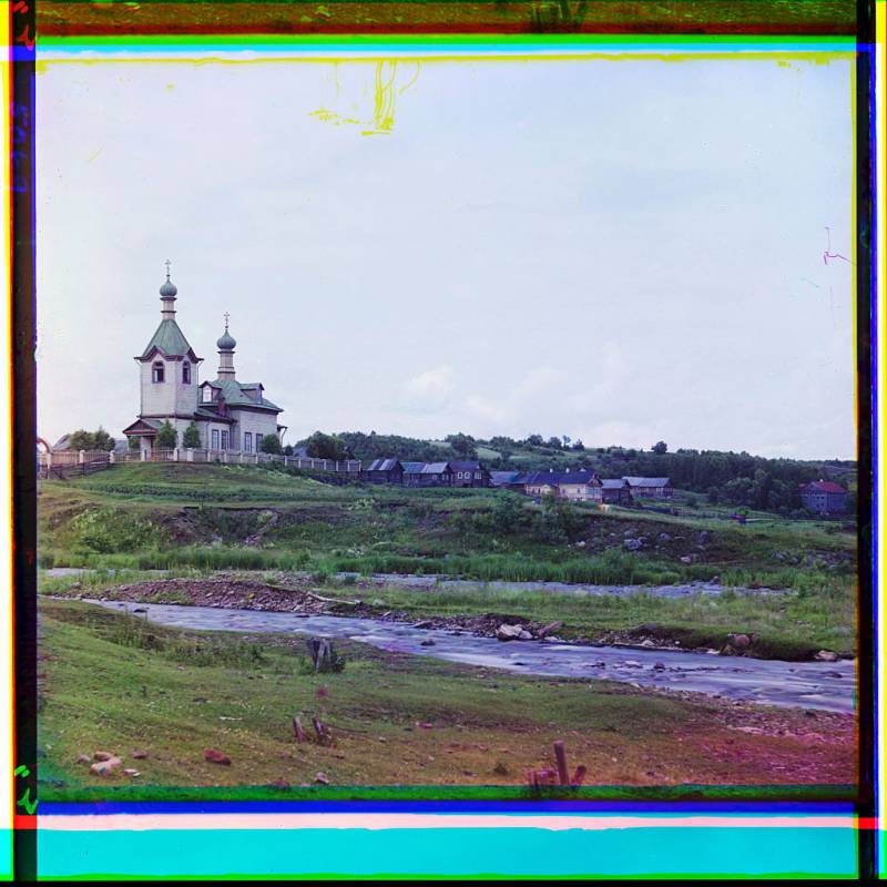 church_surrounded_by_small_wooden_buildings_stream_in_foreground.jpg