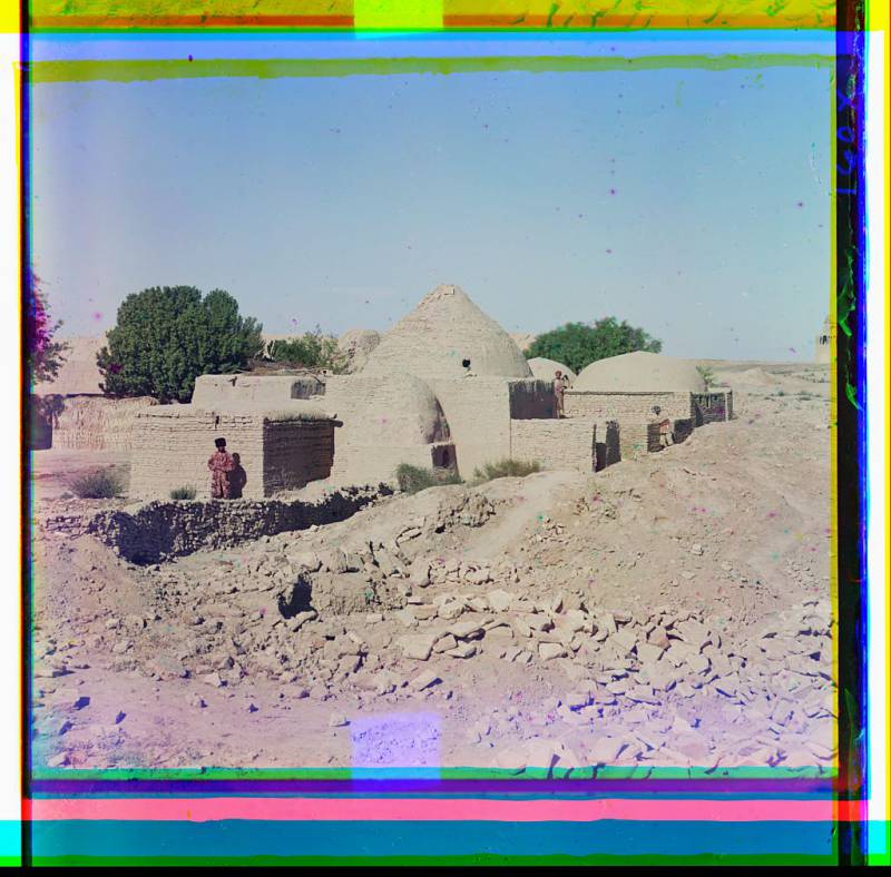 domed_adobe_buildings_with_several_people_standing_nearby_part_of_crumbling_mosque_visible_in_background_on_right.jpg