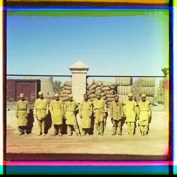 group_of_laborers_standing_posed_in_front_of_a_chain-link_fence_with_sacks_piled_behind_fence.jpg