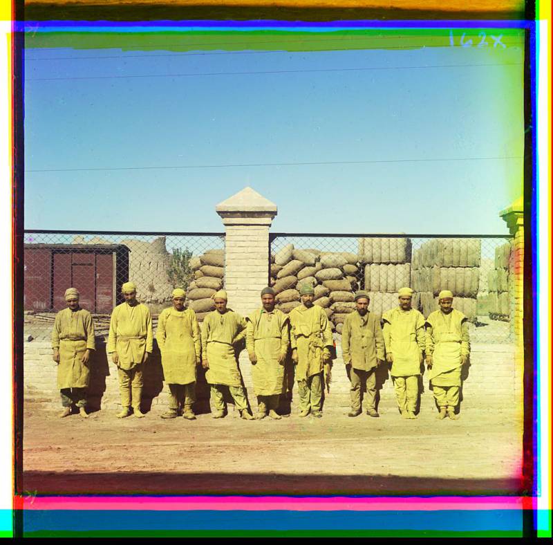 group_of_laborers_standing_posed_in_front_of_a_chain-link_fence_with_sacks_piled_behind_fence.jpg